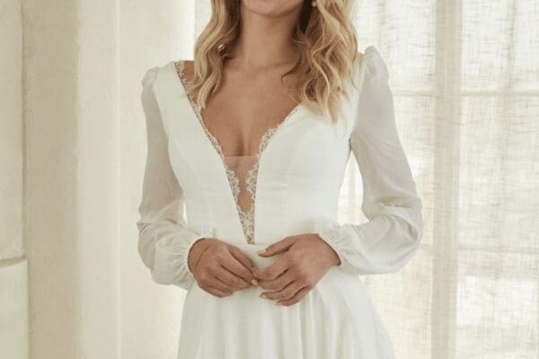 A woman in an elegant white dress with lace details and long sleeves stands in front of sheer curtains, exuding a sense of serene elegance.