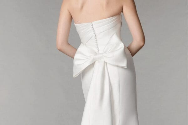 A woman in a strapless white wedding dress with a large detachable bow at the back. The dress has a row of buttons and drapes elegantly, evoking elegance.