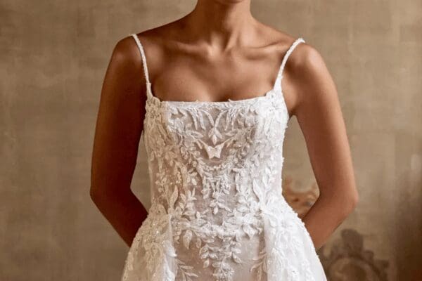 A bride in a detailed, lace wedding dress with thin straps stands and square neckline against a soft, neutral background. The intricate floral patterns convey elegance and grace.