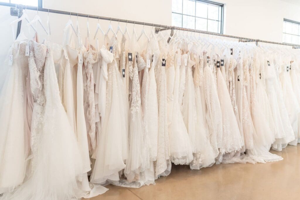 A rack of elegant white and ivory wedding dresses on hangers in a brightly lit boutique. The dresses showcase various styles and lace patterns.