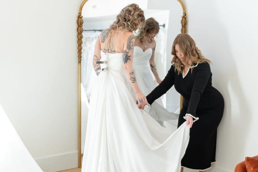 Bride trying on a wedding dress while a bridal stylist adjusts the gown in front of a mirror at a bridal boutique.