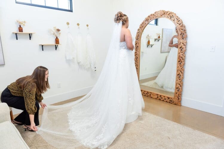 Bride trying on a wedding dress with a long train while a stylist adjusts the fabric in a bridal boutique fitting room.