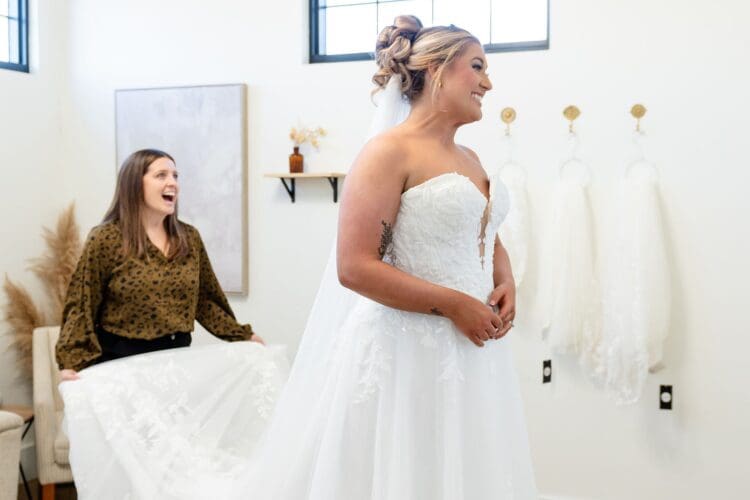 Bride smiling in a strapless lace wedding dress while a stylist lifts the train during a bridal fitting appointment.