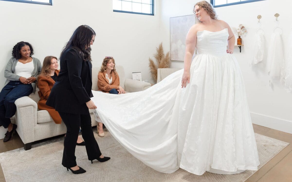 A bride tries on a flowing white wedding dress in a bright room. A stylist assists, while four friends watch, smiling from a couch. The mood is joyful.