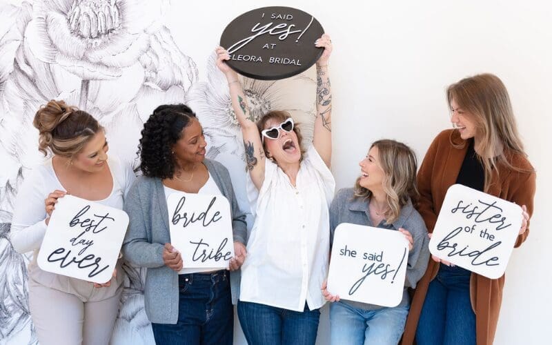 A joyful group of five women holds celebratory signs about a wedding. The central woman raises a sign overhead with excitement. Casual, happy atmosphere.