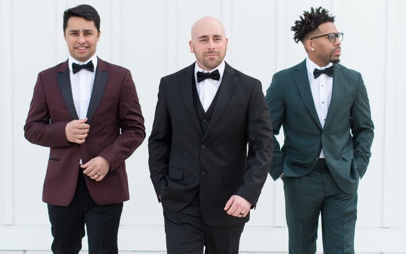 Three grooms in formal tuxedos walking outdoors at a wedding venue