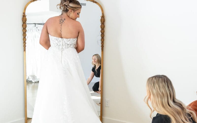 Bride standing in front of a mirror during a wedding dress fitting, wearing a strapless lace A-line gown with a long train.