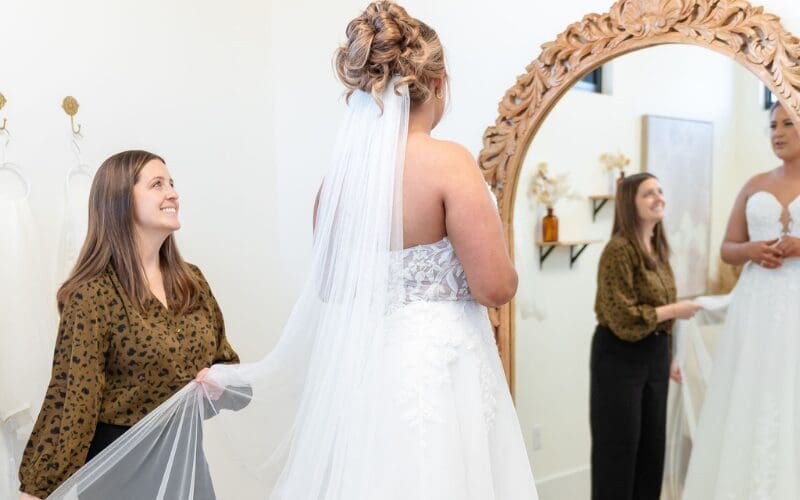 Bride in a wedding dress during a fitting while a stylist adjusts her veil in a bridal boutique