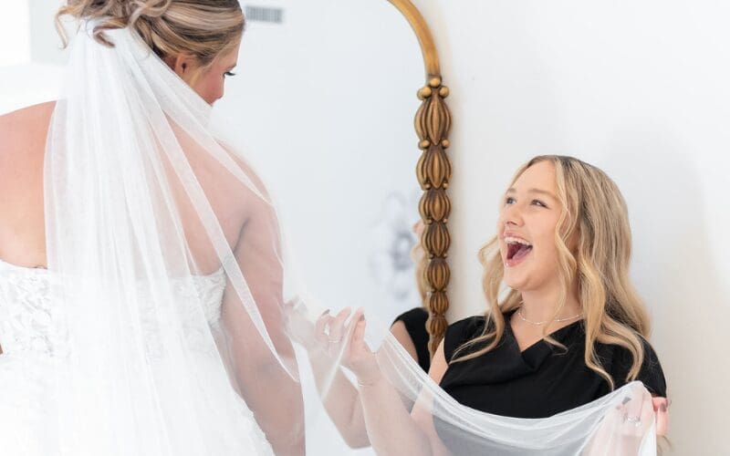 Bride smiling during a wedding dress fitting while stylist adjusts her veil