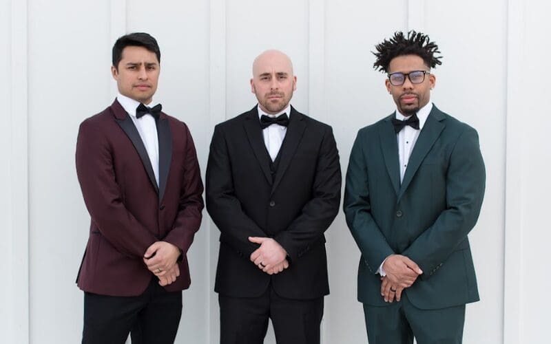 Three grooms stand side by side wearing formal wedding attire, showcasing different tuxedo and suit color options.