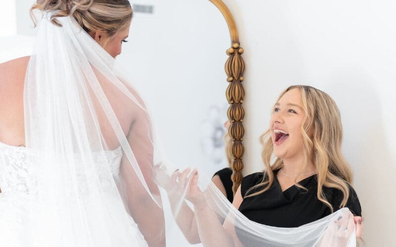 Bride wearing a wedding veil during a wedding dress fitting while a bridal consultant helps with styling details.