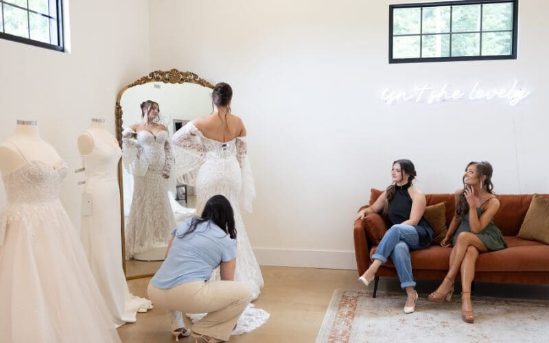 Bride trying on a lace wedding dress while her stylist adjusts the gown and friends watch
